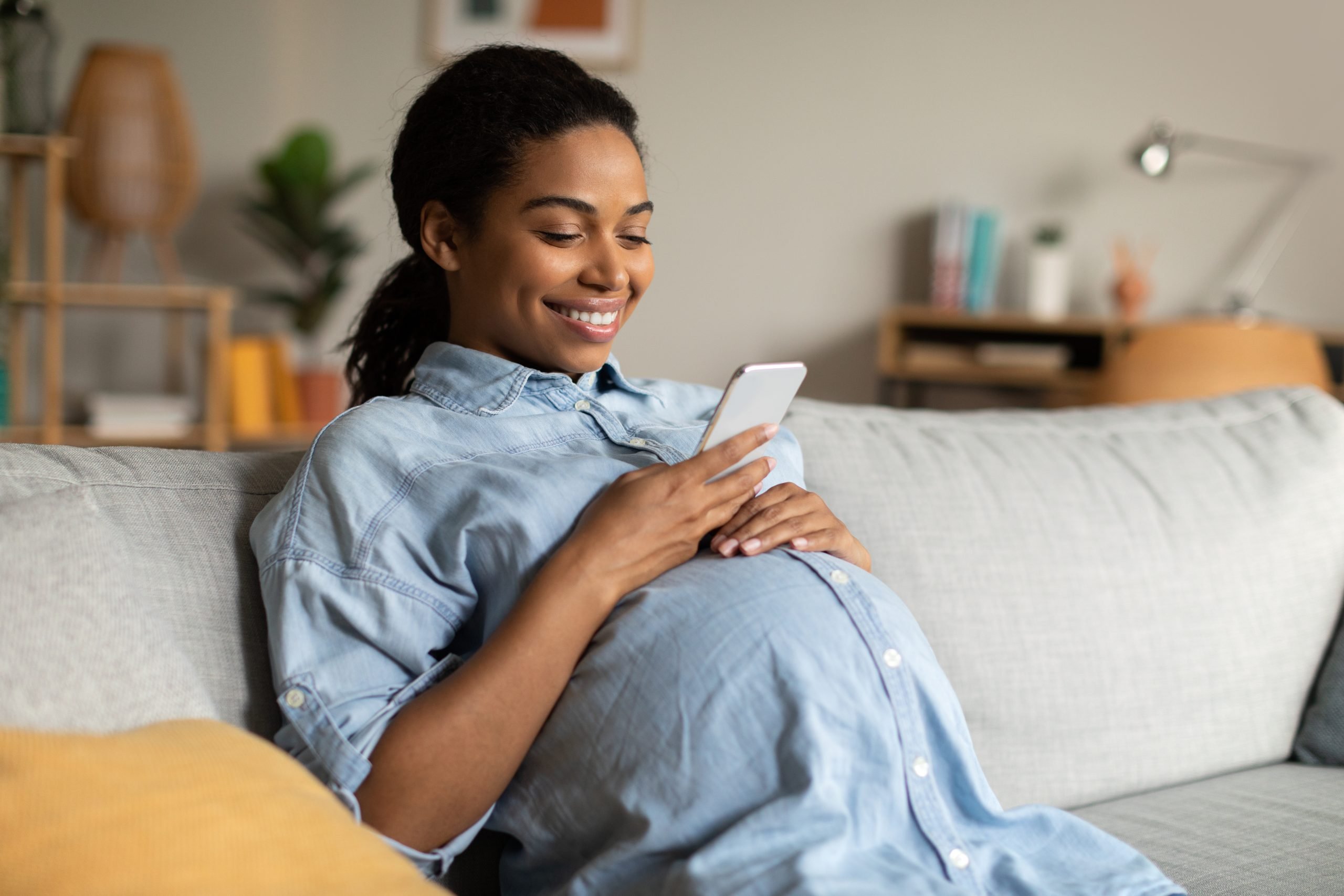 Mulher grávida sentada no sofá sorrindo e olhando celular